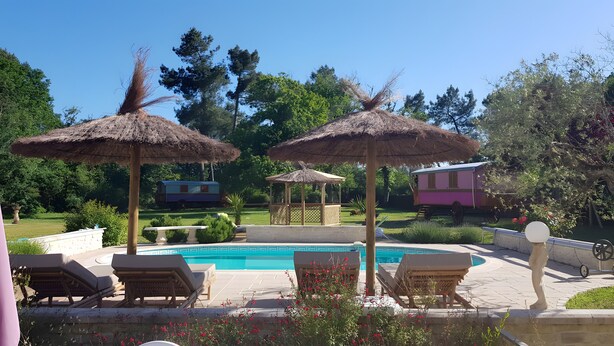 Zona de piscina con sillones de descanso, sombrillas de paja y un edificio pequeño al fondo.