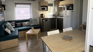 A compact kitchen with a microwave, stove, and refrigerator. There is a dining table with a book on it and a sofa with cushions. A window provides natural light. - Mobile Home 'Mobil Home Arès' in Arès, Near the Beach and Arcachon Bay (Arès)