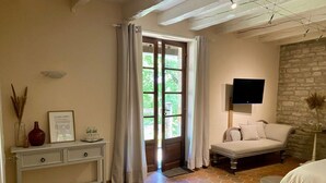 A room with a hexagonal tiled floor, a white sofa, a television mounted on a stone wall, and a door leading outside. - Quercy Room – Romantic & Authentic Retreat (Gramat)