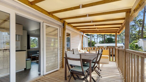 A covered deck with a table and chairs, a sliding glass door leading to a kitchen, and a view of the surrounding area. - Bungalow "Les Lodges Des Pins" with Shared Pool (Lège-Cap-Ferret)