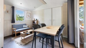 A modern living room with a sofa, dining table, chairs, and a view through a window. - Bungalow "Les Lodges Des Pins" with Shared Pool (Lège-Cap-Ferret)