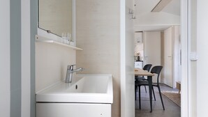 Een moderne badkamer met een wit wastafelmeubel, spiegel en plank. In de achtergrond is een zithoek met een tafel en stoelen te zien.