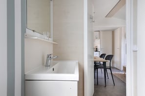 Un baño moderno con un lavabo blanco, espejo y estante. Se ve un comedor con una mesa y sillas al fondo.