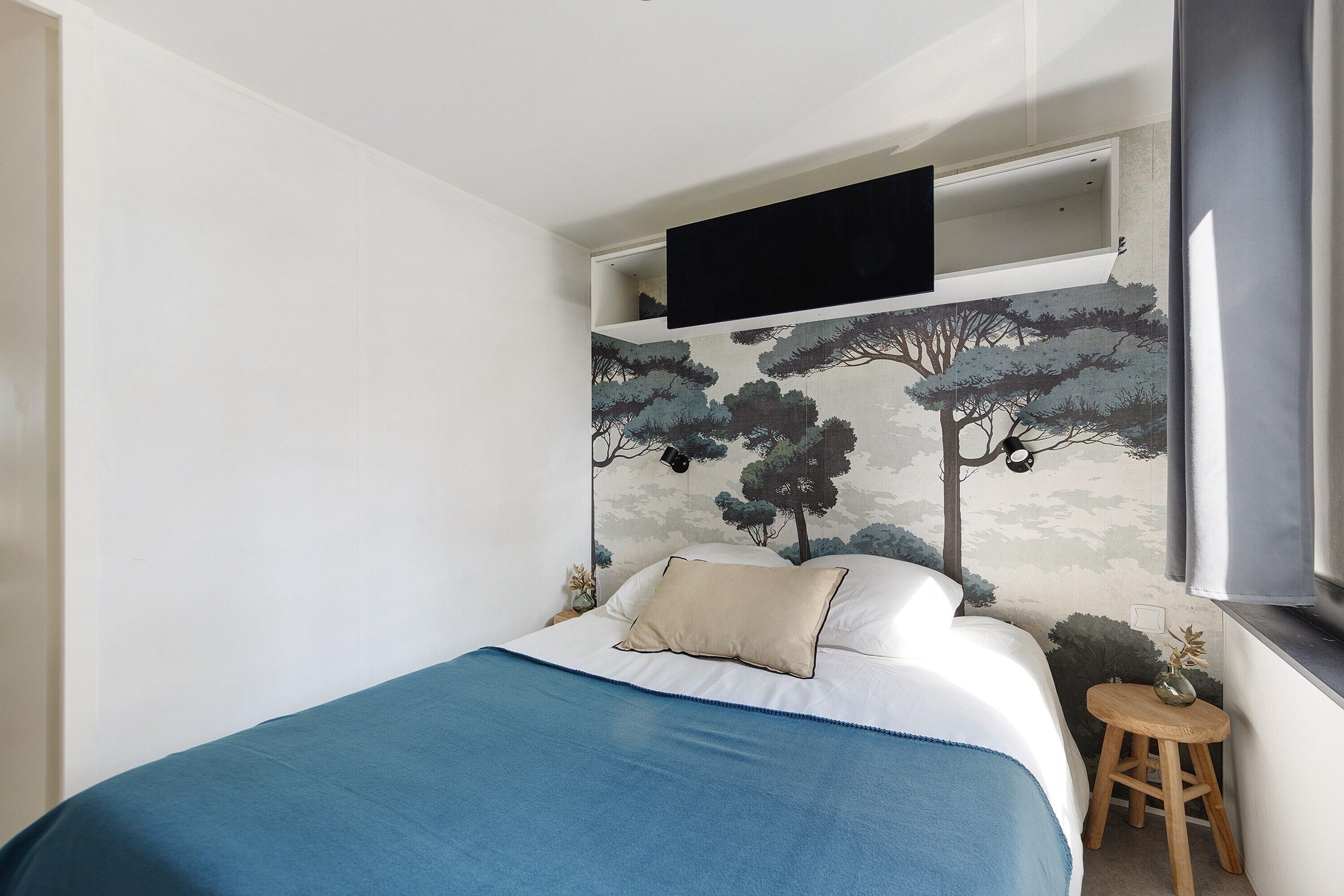 A bedroom with a bed, a mounted TV, a mural, and a small stool with a plant.