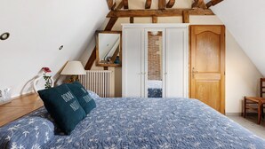 Quarto com teto inclinado, cama com colcha florida azul, cadeira de madeira, porta de madeira e radiador.