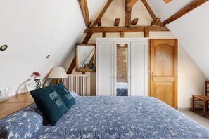 A bedroom with a sloped ceiling, a bed with a blue floral bedspread, a wooden chair, a wooden door, and a radiator. - Gîte Roses De Loire with Private Terrace and Wi-Fi in Bréhémont (Bréhémont)
