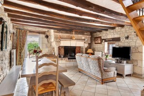 A rustic living room with exposed wooden beams, a stone fireplace, a sofa, a TV, and a dining area with a table and chairs. - Gîte Roses De Loire with Private Terrace and Wi-Fi in Bréhémont (Bréhémont)