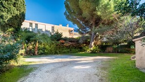 Une allée de gravier mène à un bâtiment à l’architecture moderne, entouré d’une végétation luxuriante et d’arbres.