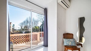Une pièce dotée d’une porte-fenêtre coulissante donnant sur un balcon extérieur, d’une chaise en bois avec un coussin à motifs et d’un climatiseur fixé au mur.