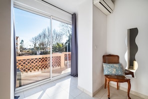 A room with a sliding glass door leading to an outdoor balcony, a wooden chair with a patterned cushion, and a wall-mounted air conditioning unit. - Courtille sur Mer – Sea View and Serenity in Fréjus (Fréjus)