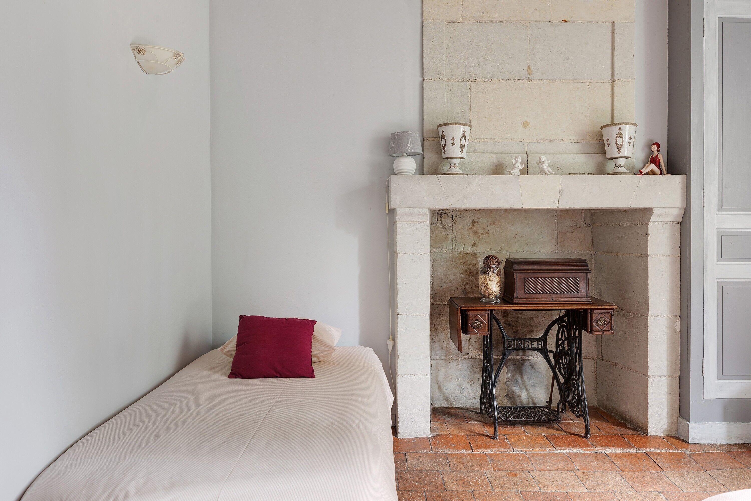 Een slaapkamer met een bed, een rood kussen, een naaimachine en een haardmantel met decoratieve objecten.