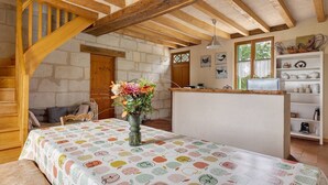 Een rustieke kamer met zichtbare houten balken, een tafel met een tafelkleed met fruitprint, een vaas met bloemen en een keukenhoek met planken.