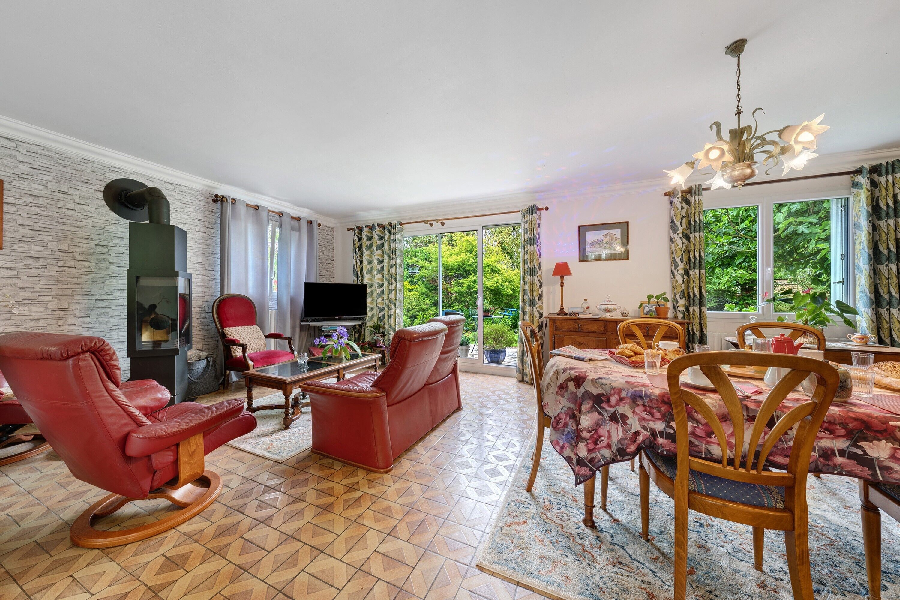 Een woonkamer met een open haard, een televisie, een eethoek met een tafel en stoelen, en uitzicht op groen buiten.