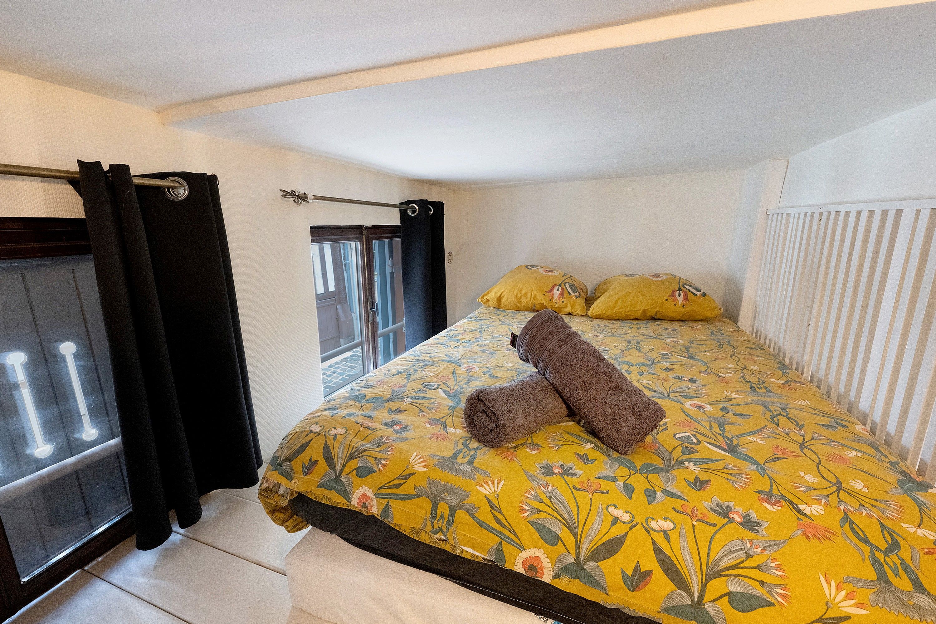 Quarto com cama, edredom floral, duas almofadas, uma toalha, janela com cortinas e radiador.