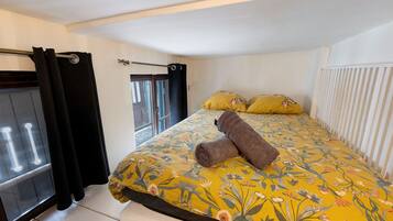 Quarto com cama, edredom floral, duas almofadas, uma toalha, janela com cortinas e radiador.