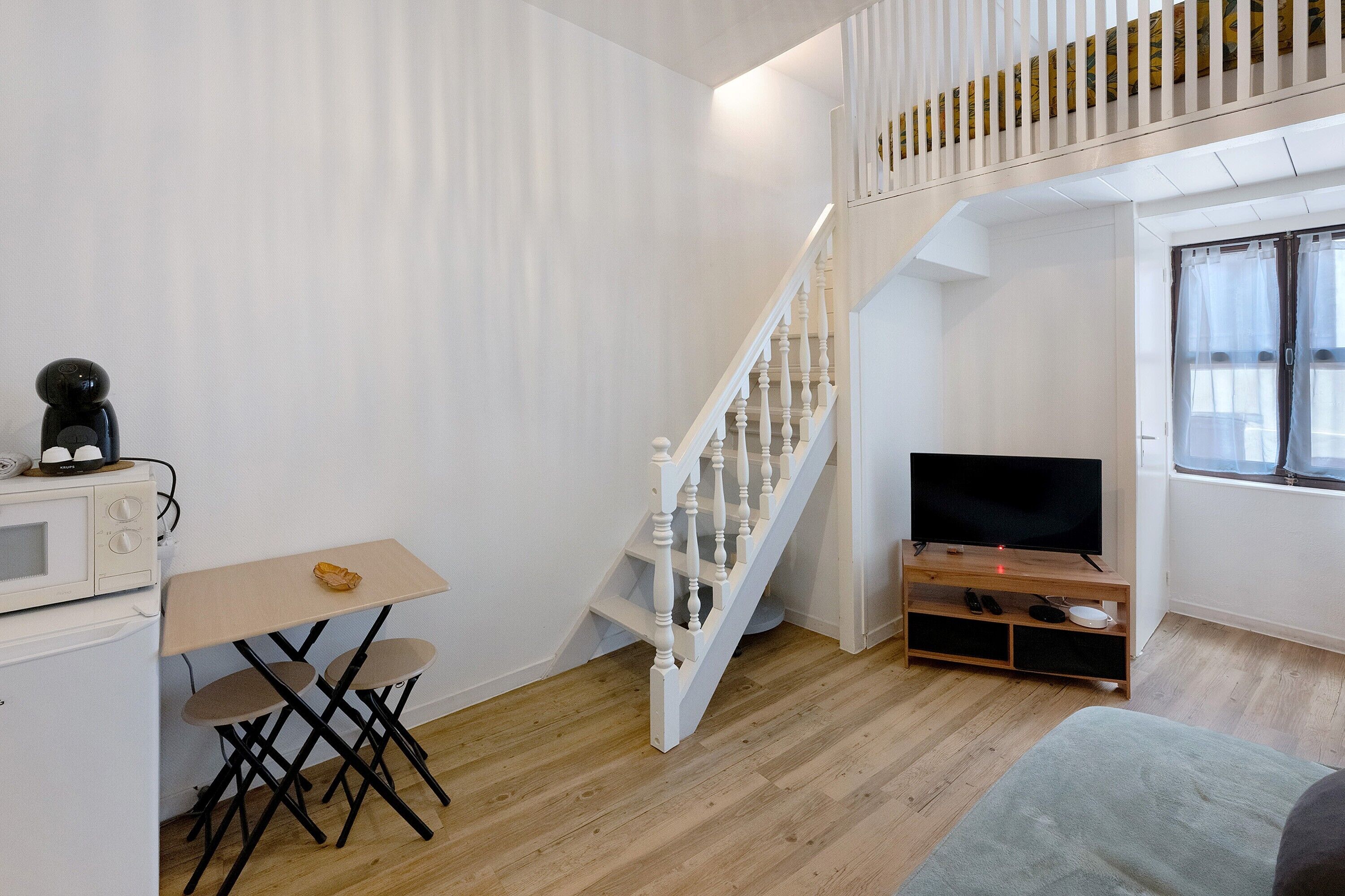 Un espace de vie compact doté d’un plancher en bois, d’un escalier blanc, d’un téléviseur à écran plat, d’une petite table et d’un four à micro-ondes.