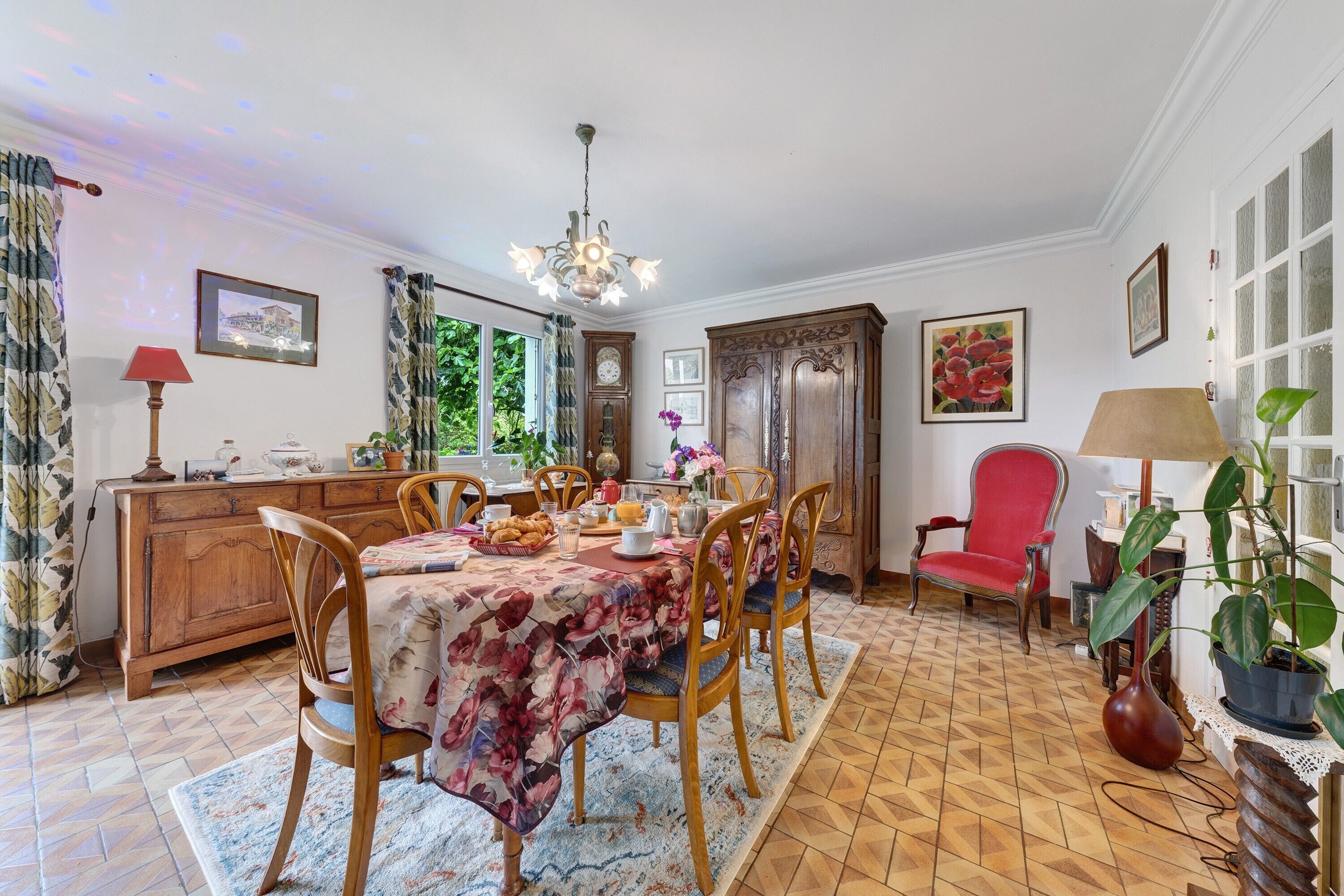 Un comedor con una mesa dispuesta para cuatro personas, rodeada de sillas de madera, una araña y un sillón rojo.