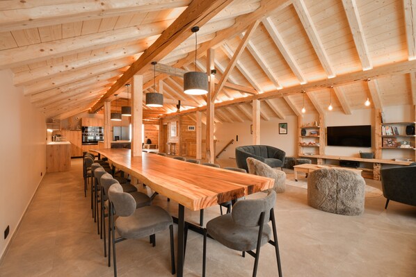 Een ruime loft met hoge plafonds, houten balken, een lange eettafel, meerdere stoelen, een bank en een televisie.