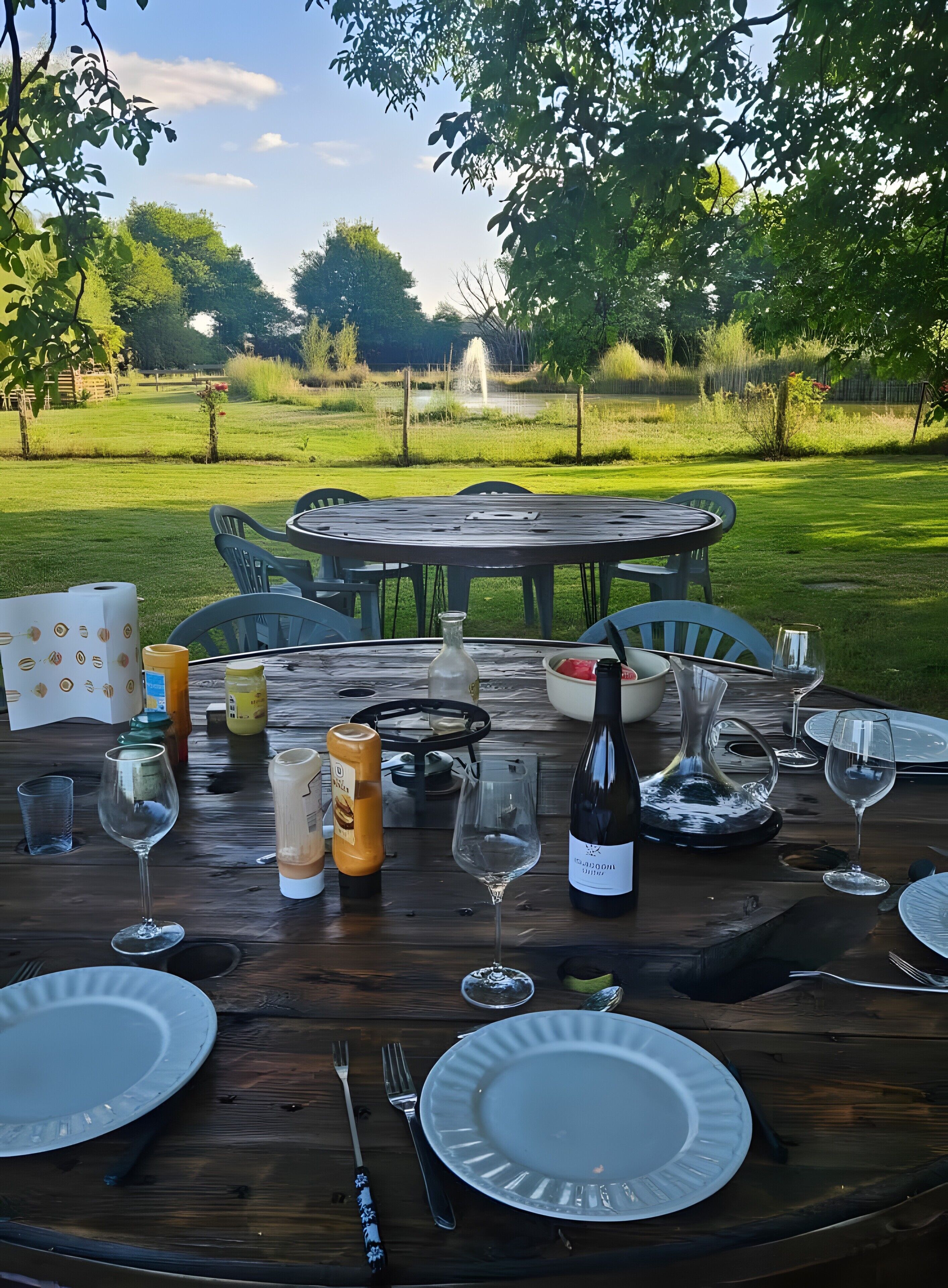 Een buitentafel gedekt met borden, bestek en een fles wijn, omringd door stoelen en uitzicht op een grasveld.