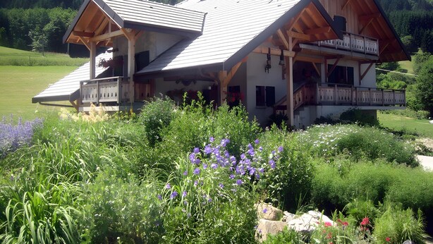 Une maison à deux étages dotée d’un balcon, entourée d’une végétation luxuriante et de fleurs.