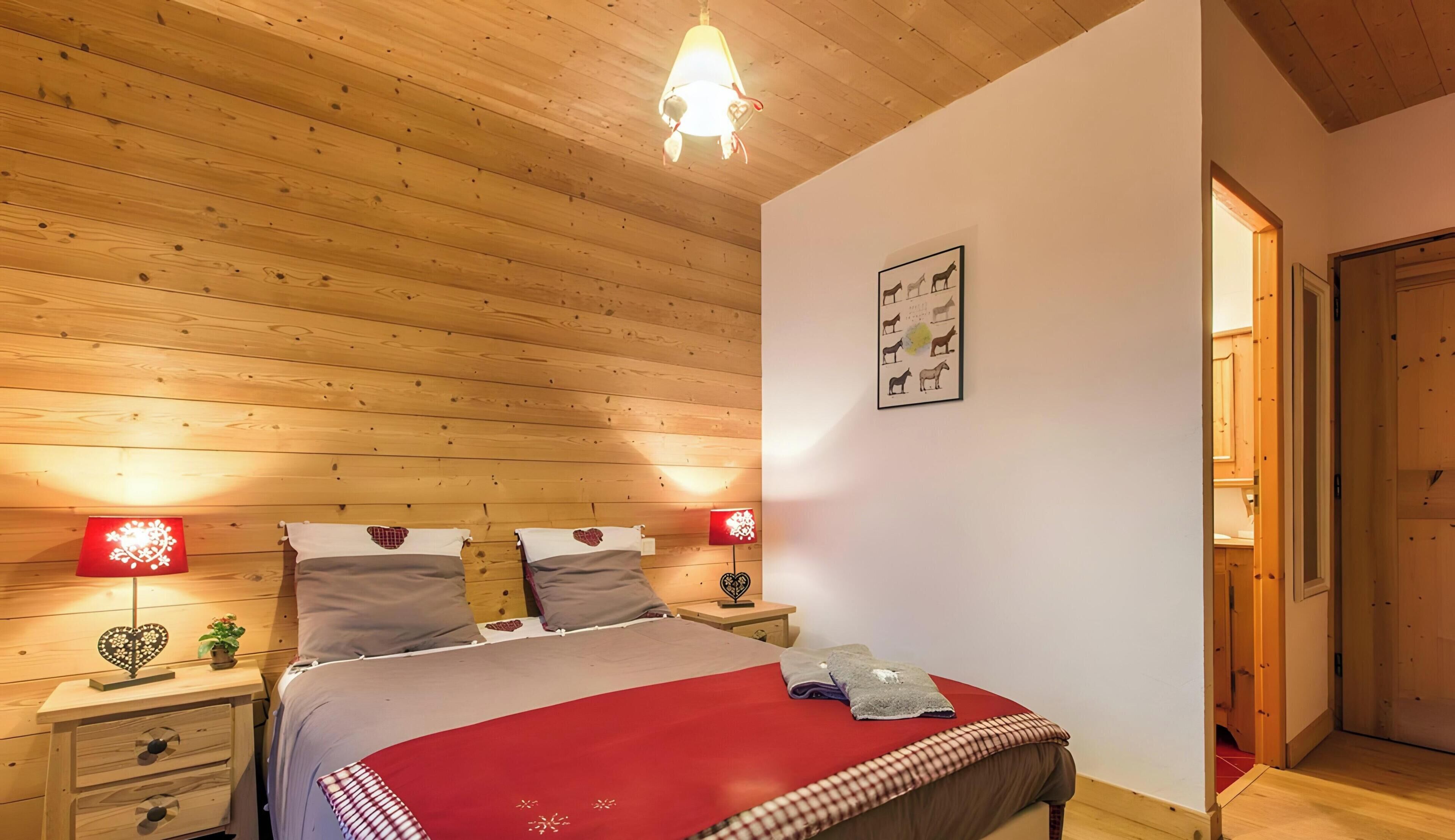 Un dormitorio con techo de madera, dos mesitas de noche con lámparas, una cama con colcha roja y una puerta que da a otra habitación.