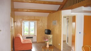 Ein Zimmer mit Holzboden, einer Couch, einem kleinen Tisch mit einer Vase, einem Fernseher und einem Fenster mit Blick auf Grünflächen.