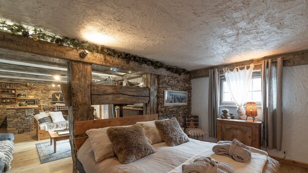 Un dormitorio rústico con pared de piedra, vigas de madera, una cama con almohadas, una mesita de noche y una ventana con cortinas.