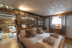 A rustic bedroom with a stone wall, wooden beams, a bed with pillows, a nightstand, and a window with curtains. - Apartment in Authentic Chalet with Mountain View, Shared Pool & Wi-Fi (Les Contamines-Montjoie)