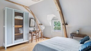 Un dormitorio con techo inclinado, una cama con ropa de cama azul, un escritorio de madera, una silla, un armario y una ventana.