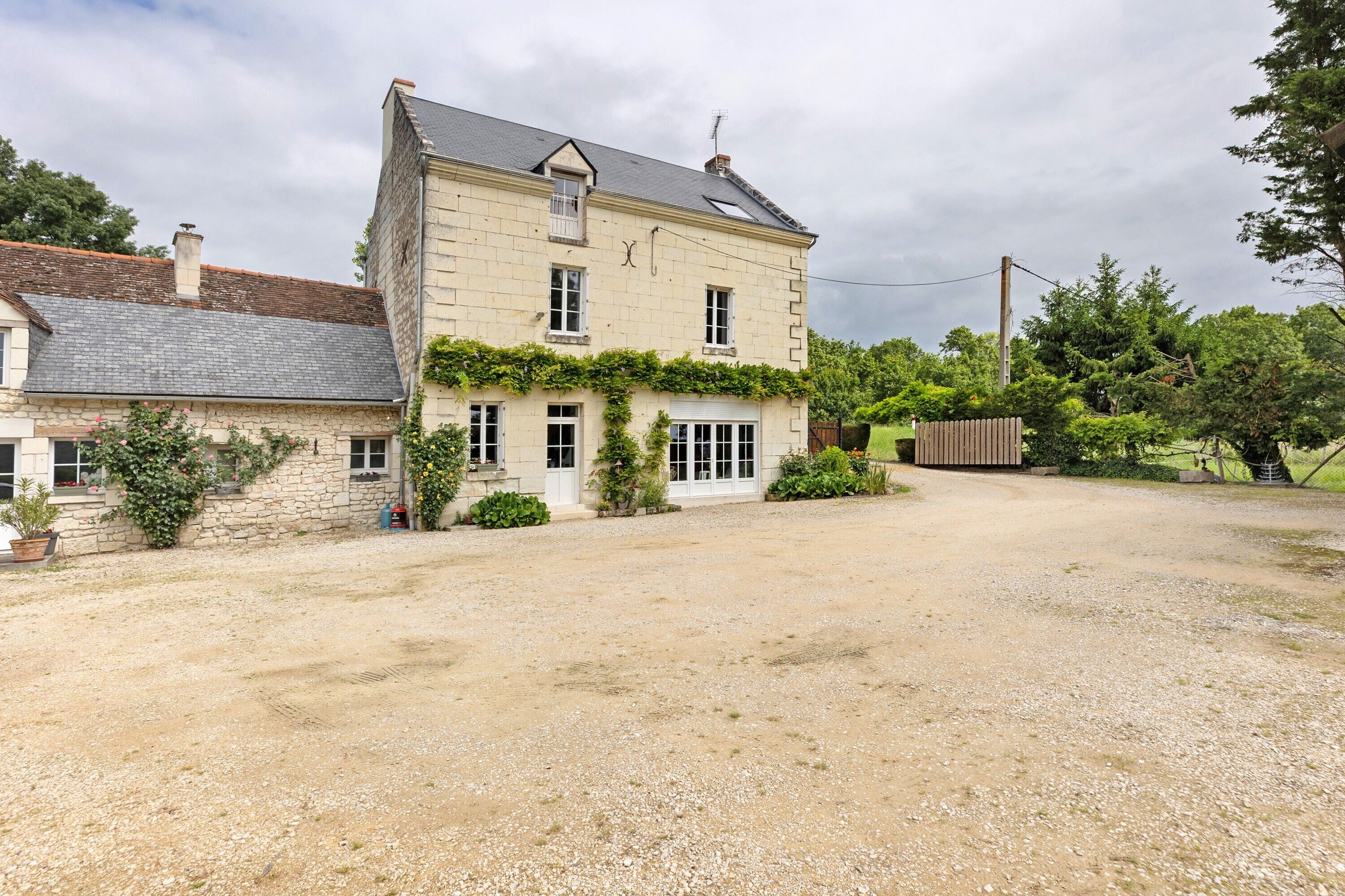 Une maison en pierre à deux étages, avec une allée en gravier, entourée de verdure et d’arbres.