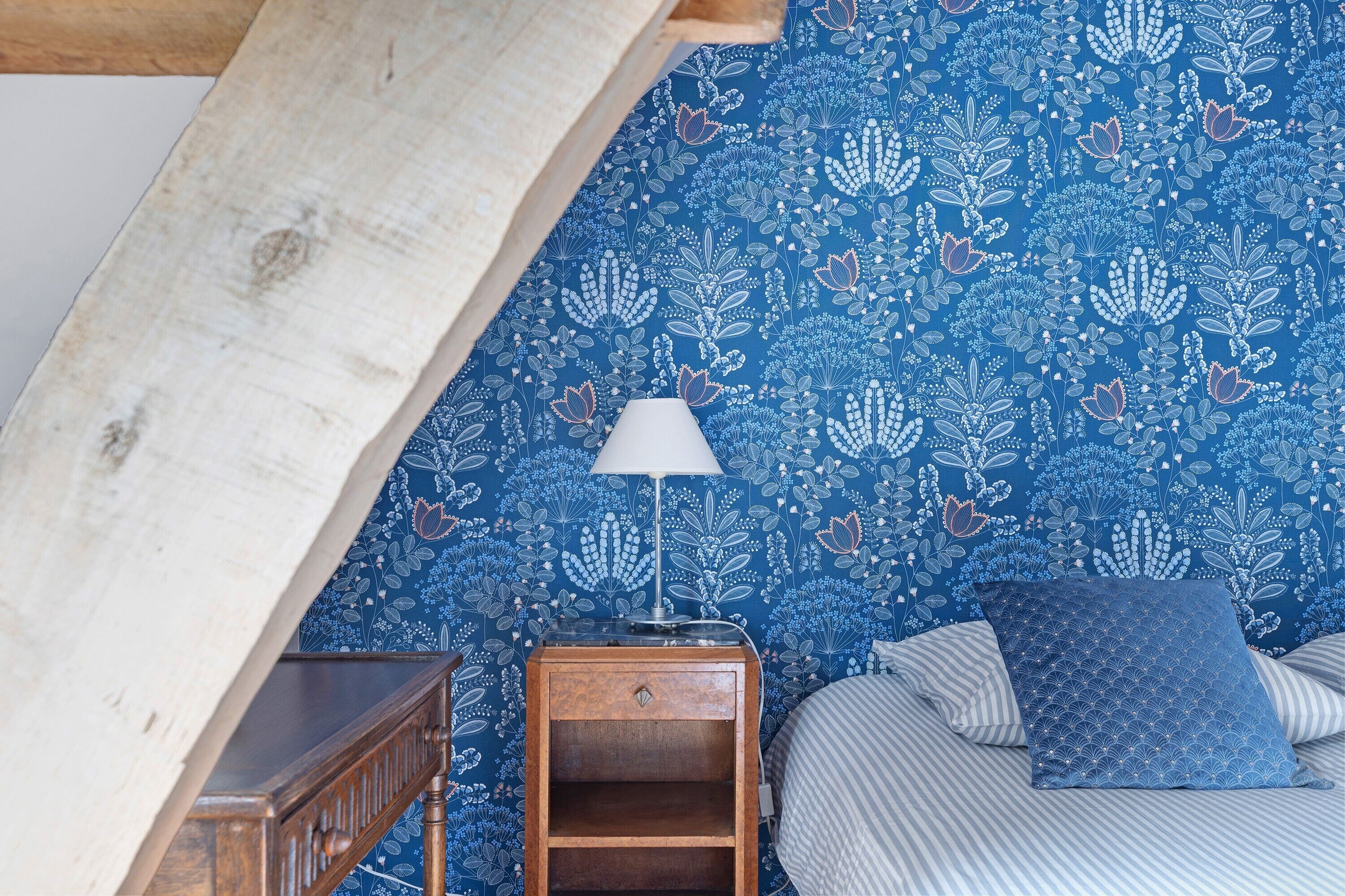 Een slaapkamer met blauw bloemenbehang, een houten nachtkastje met een lamp, een bed met gestreept beddengoed en een schuine plafond.