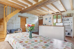 Une pièce rustique avec des poutres apparentes, un lit recouvert d’une couvre-lit à motifs, une chaise en bois, un vase de fleurs et un coin cuisine avec des étagères.