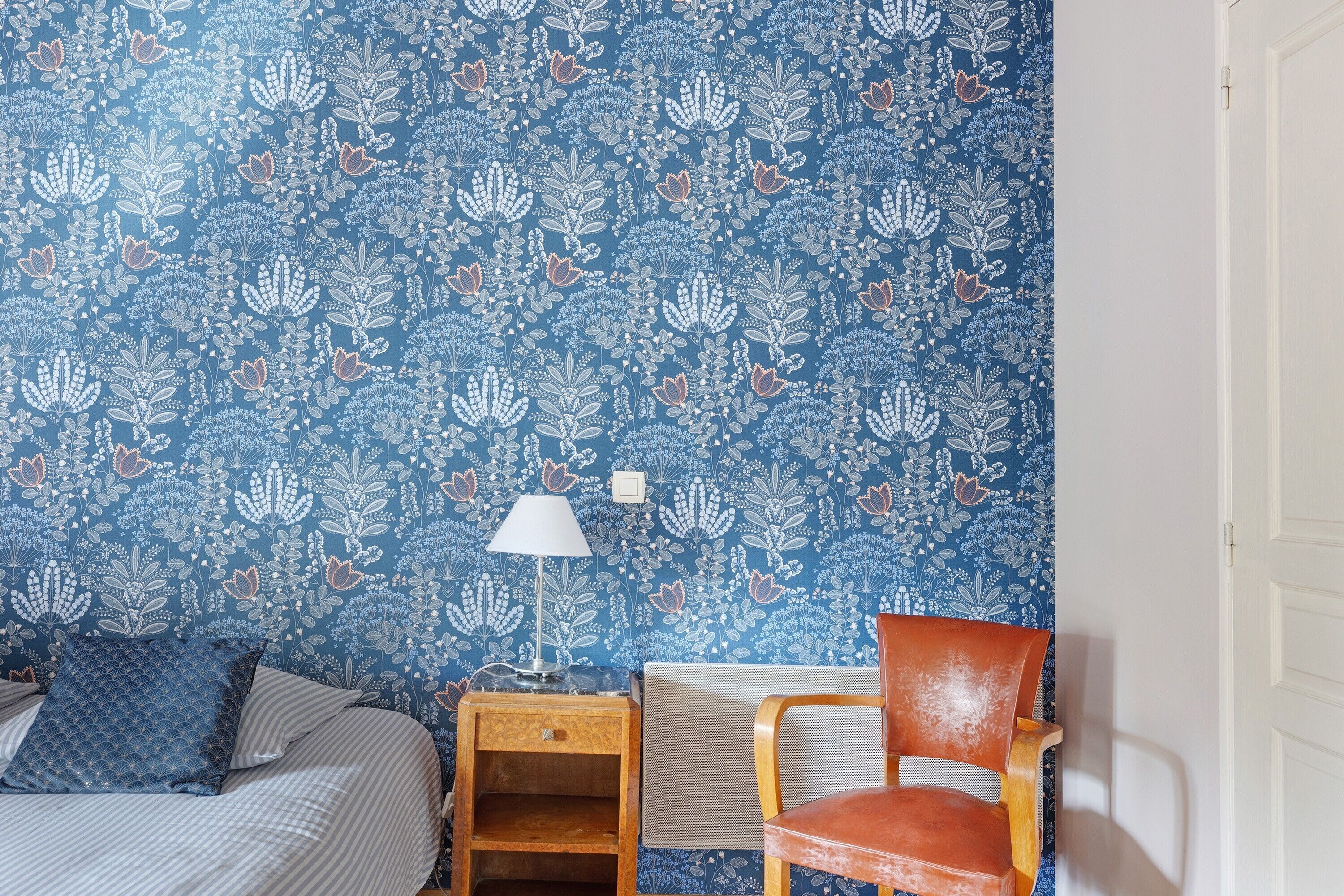 Una camera da letto con carta da parati a motivi floreali, un letto con biancheria a righe, un comodino in legno con una lampada e una sedia rossa.