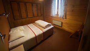 Una habitación de cabaña de madera con una cama, una ventana con cortinas, una lámpara en el techo y un radiador.