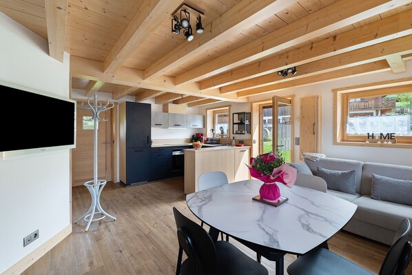 A modern interior with a dining area, kitchen, and living space, featuring wooden beams, a flat-screen TV, and a sofa.