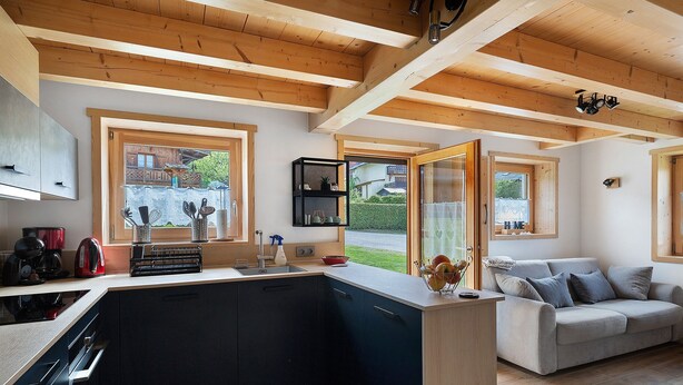 A modern kitchen with wooden beams, a grey sofa, and a view of a house through large windows.