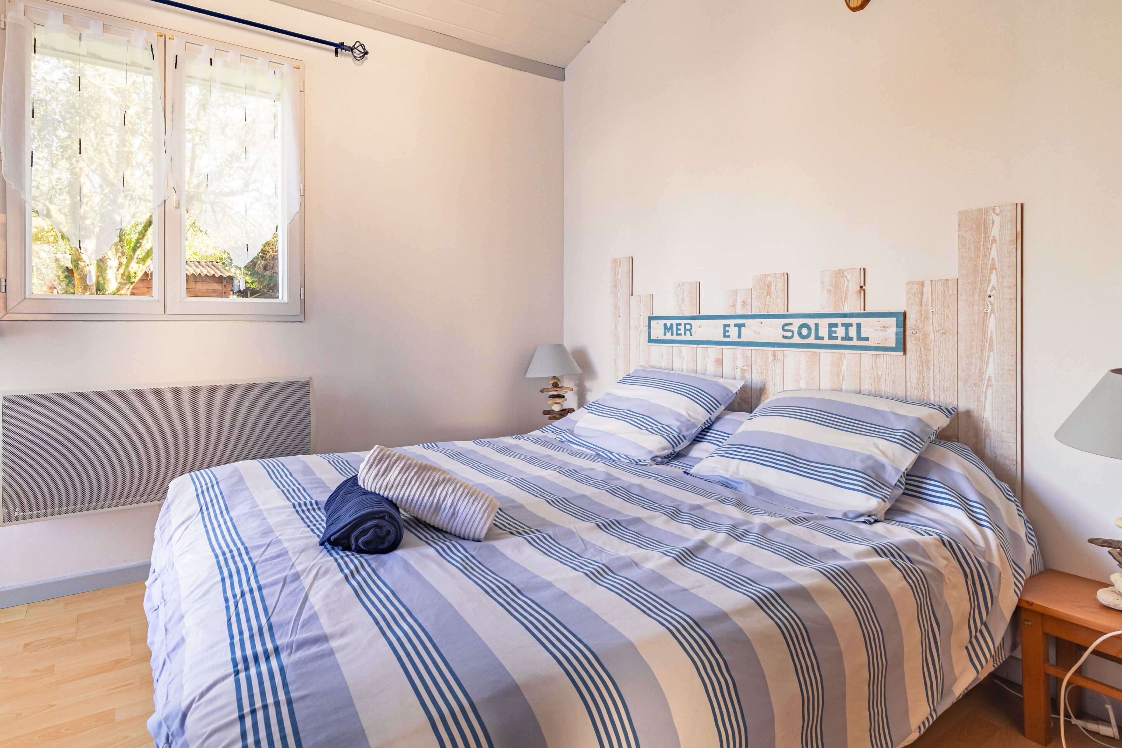 Un dormitorio con cama, ventana, cartel en la pared, mesita de noche y lámpara.