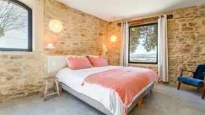A bedroom with a stone wall, a bed with pink bedding, a wooden stool, a blue chair, and a view of the outdoors. - Holiday Home - Les Gîtes De Figon - Private Pool, Wi-Fi, Air Conditioning (Laudun)