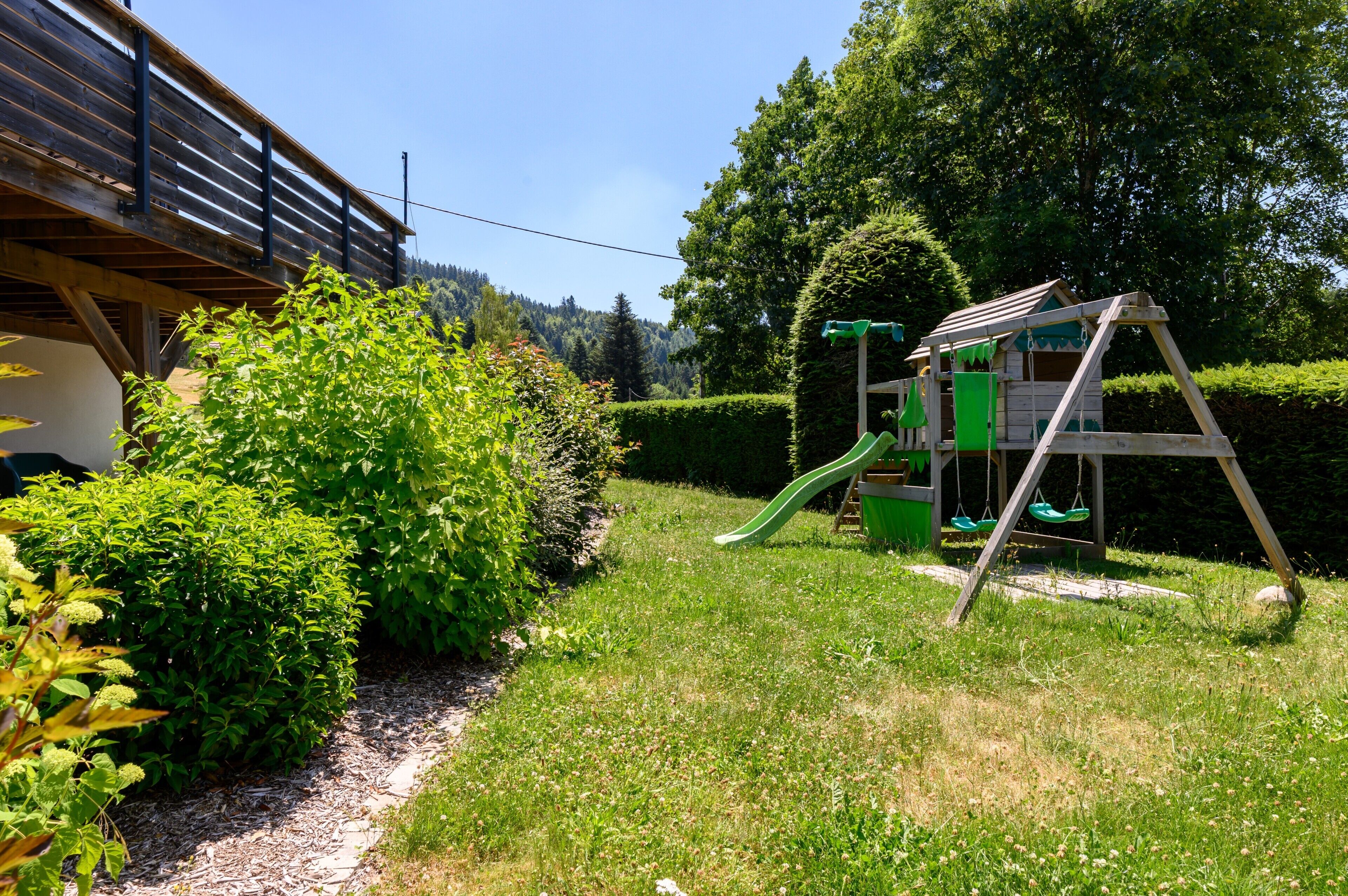 A wooden play structure with a slide, surrounded by well-maintained hedges and a clear sky.