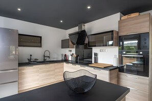 A modern kitchen with a black dining table, wooden cabinets, a built-in oven, and a large window. - Gîte Perce Neige: Spa, Gym, Near Gérardmer and Ski Slopes (Gerbépal)