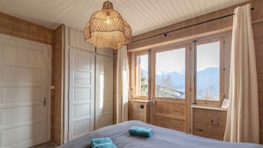 Quarto com cama, duas toalhas, luminária pendente e vista para montanhas por grandes janelas.