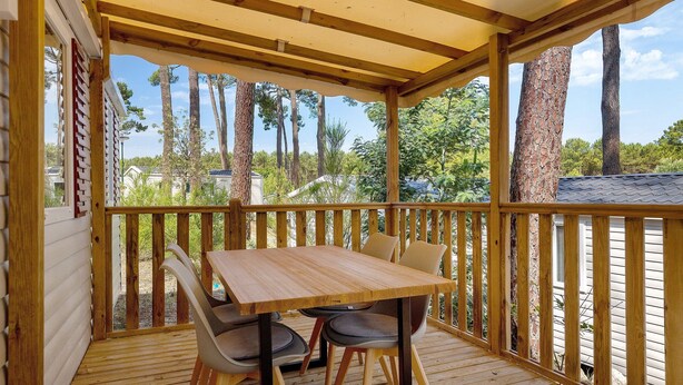 Un espace extérieur abrité, doté d’une table et de chaises en bois, entouré d’une balustrade en bois, et offrant une vue sur un quartier résidentiel avec des arbres et des maisons.