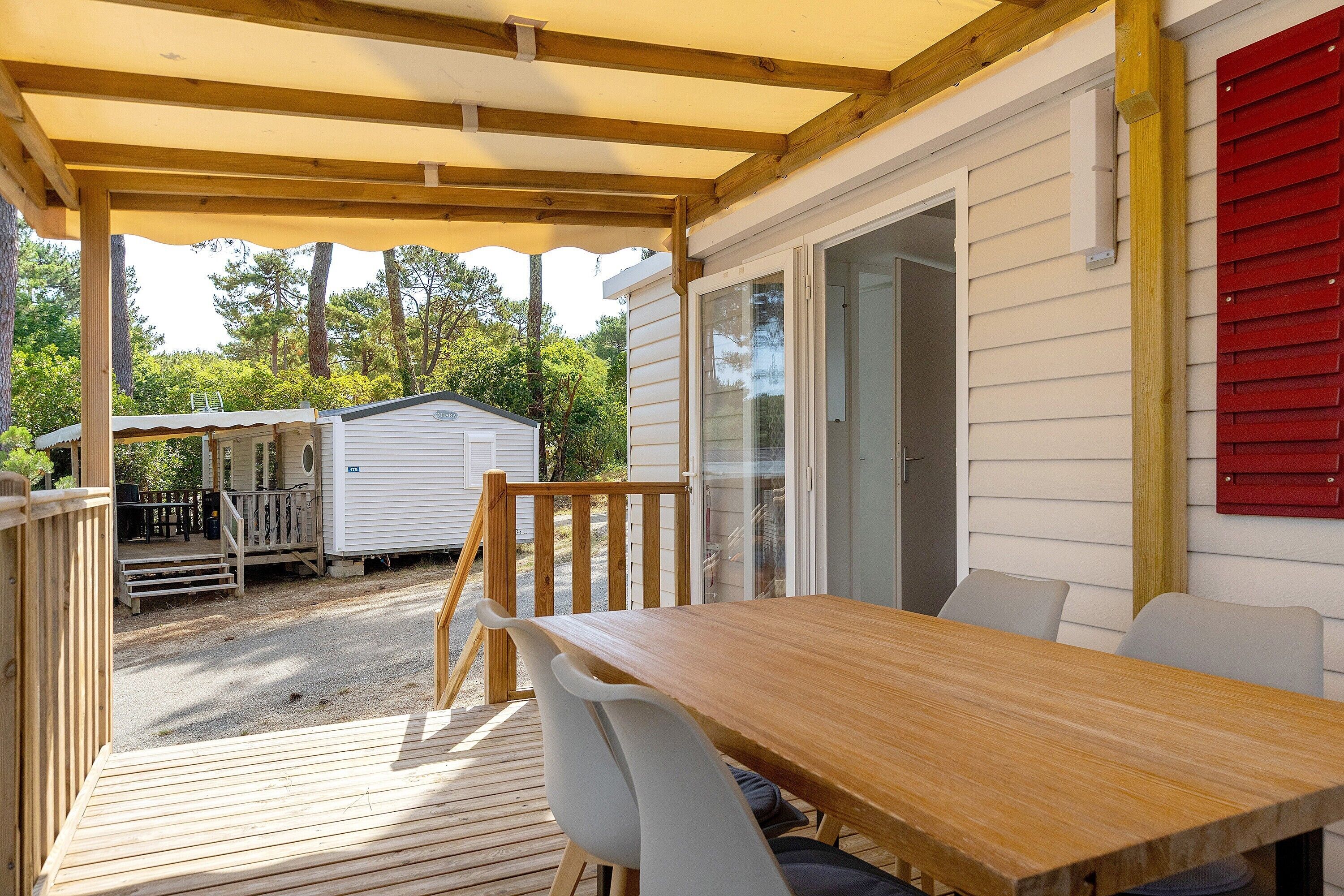 Une terrasse couverte avec une table et des chaises en bois, une plateforme et une vue sur une maison avec une porte rouge.