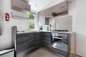 A modern kitchen with dark cabinetry, a gas stove, an oven, and a window with a view of greenery. - Bungalow "Les Lodges Des Pins" – Cozy Retreat in Lège-Cap-Ferret (Lège-Cap-Ferret)
