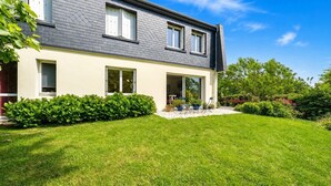 Ein modernes, zweistöckiges Haus mit gepflegtem Rasen, einer Terrasse mit Gartenmöbeln und verschiedenen Pflanzen und Sträuchern.
