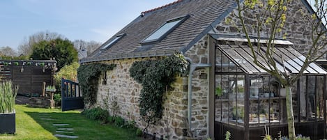 A stone house with a sloped roof, a glass greenhouse, and a well-maintained garden.
