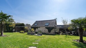 Une maison en pierre avec un toit en pente, une cour verdoyante et un chemin de pierre menant à l’entrée.