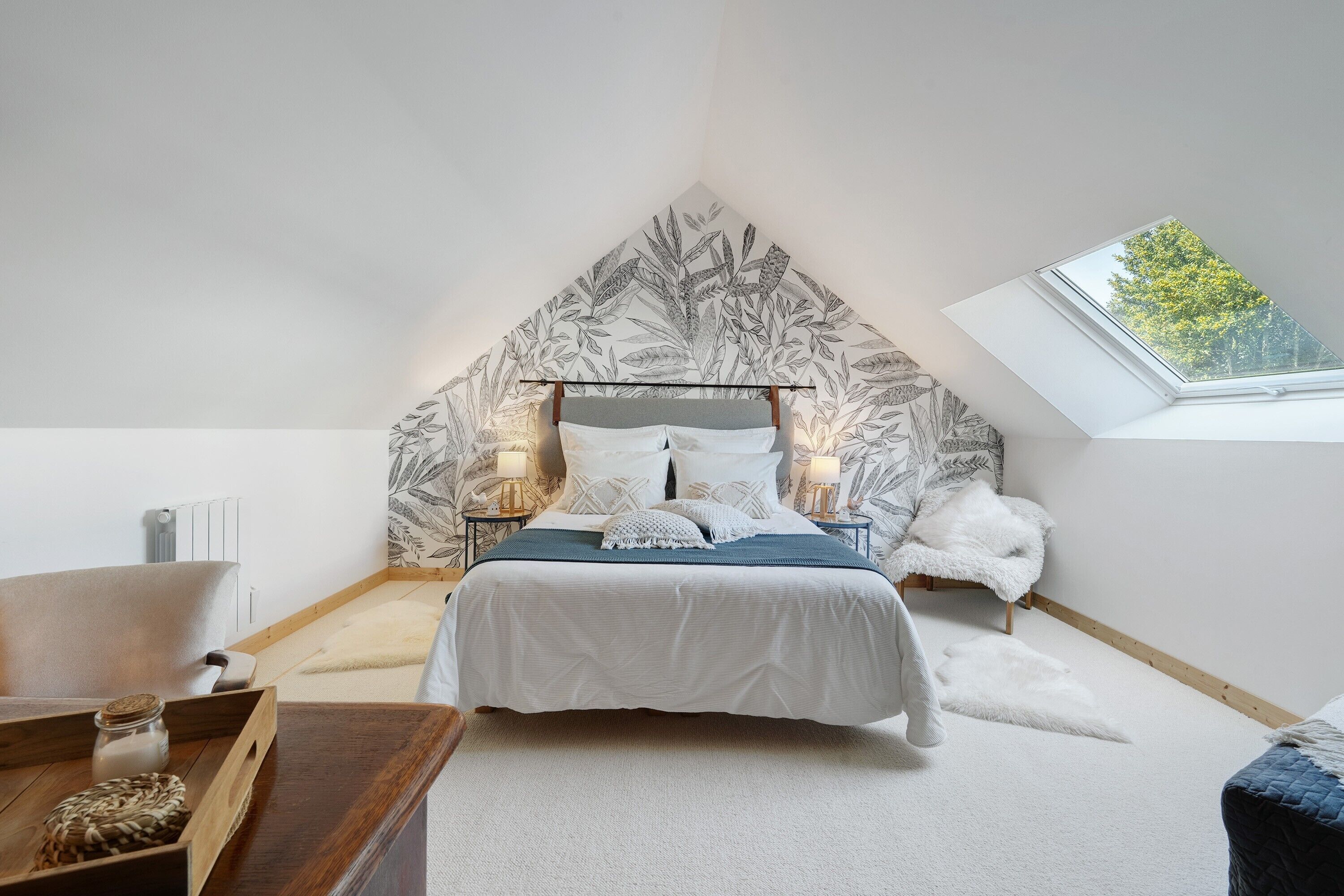 Une chambre avec un plafond en pente, un puits de lumière, un lit avec une literie blanche, une table de chevet en bois et un fauteuil blanc moelleux.