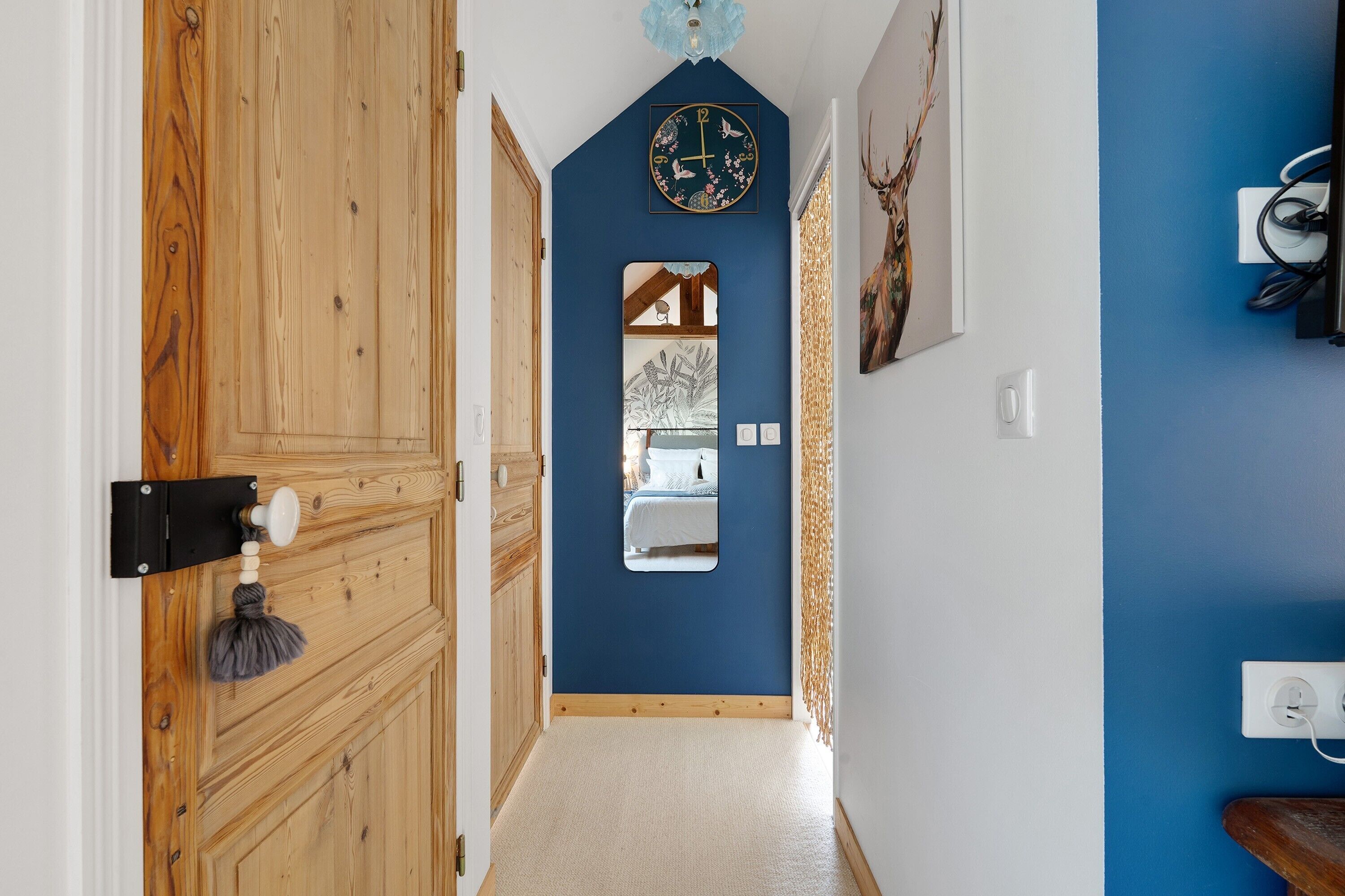 Ein Flur mit einer Holztür, einer blauen Wand, einer Uhr, einem Spiegel und einem Bett, das im Spiegel sichtbar ist.