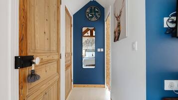 Ein Flur mit einer Holztür, einer blauen Wand, einer Uhr, einem Spiegel und einem Bett, das im Spiegel sichtbar ist.